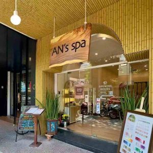 Exterior view of AN's Spa featuring a synthetic bamboo wall and ceiling that creates a strong Southeast Asian resort ambiance. The natural wooden sign with "AN's spa" lettering, lush green plants, and clear glass entrance enhance the brand's eco-friendly and relaxing wellness vibe.