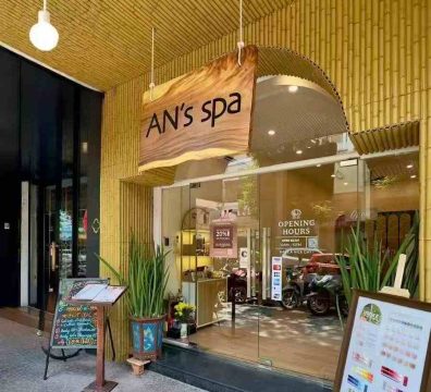 Exterior view of AN's Spa featuring a synthetic bamboo wall and ceiling that creates a strong Southeast Asian resort ambiance. The natural wooden sign with "AN's spa" lettering, lush green plants, and clear glass entrance enhance the brand's eco-friendly and relaxing wellness vibe.