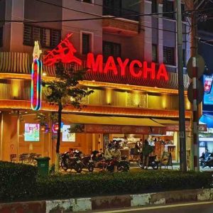 Night view of a Vietnamese street restaurant with a synthetic bamboo wall exterior, topped by a glowing red "MAYCHA" neon sign and silhouette icon. Warm yellow lighting, curved metal eaves, and bustling storefront convey a vibrant, retro-modern urban dining atmosphere.