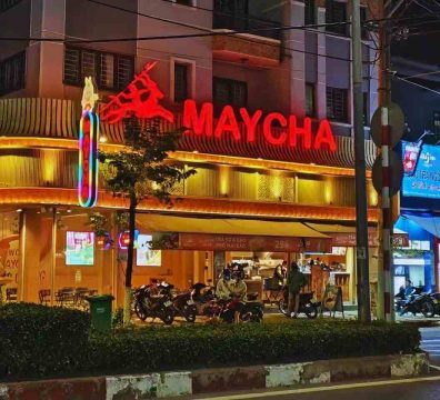 Night view of a Vietnamese street restaurant with a synthetic bamboo wall exterior, topped by a glowing red "MAYCHA" neon sign and silhouette icon. Warm yellow lighting, curved metal eaves, and bustling storefront convey a vibrant, retro-modern urban dining atmosphere.