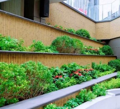 A modern urban-style terraced green courtyard incorporates synthetic bamboo elements. The scene presents a multi-level, stepped layout where dark grey metal railings and light wood-toned walls create a stark contrasting palette. Each tier is landscaped with spherical shrubs, ornamental foliage, and red flowering plants, harmoniously coexisting with the glazed building facade above.