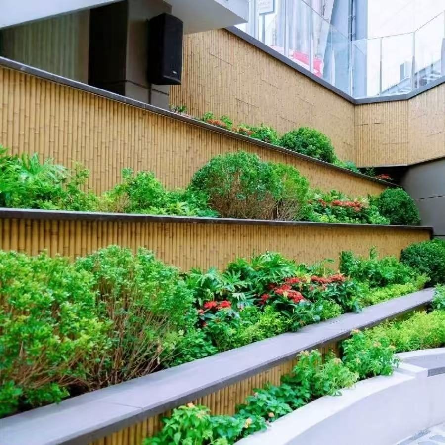 A modern urban-style terraced green courtyard incorporates synthetic bamboo elements. The scene presents a multi-level, stepped layout where dark grey metal railings and light wood-toned walls create a stark contrasting palette. Each tier is landscaped with spherical shrubs, ornamental foliage, and red flowering plants, harmoniously coexisting with the glazed building facade above.