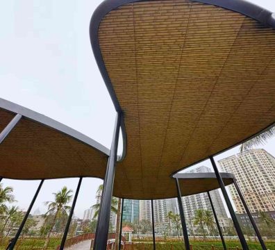 A modern outdoor shading structure features a core load-bearing framework combining multiple straight synthetic bamboo poles with curved black metal members. The poles exhibit a warm, uniform pale brown hue and a fine, consistent texture. Topped with a woven mat, the overall design masterfully blends natural materiality with contemporary aesthetics through the contrast of rigid and soft elements.