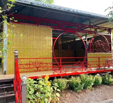 A distinctive elevated pavilion marrying rustic Southeast Asian charm with Chinese accents. Its walls are sheathed in warm synthetic bamboo poles, set on a wooden deck under a corrugated metal roof. The eye is drawn to vibrant red ironwork detailing, including arches and traditional patterns, which contrasts beautifully with the natural setting of sunflowers and shrubs, forming a cheerful and unique getaway.