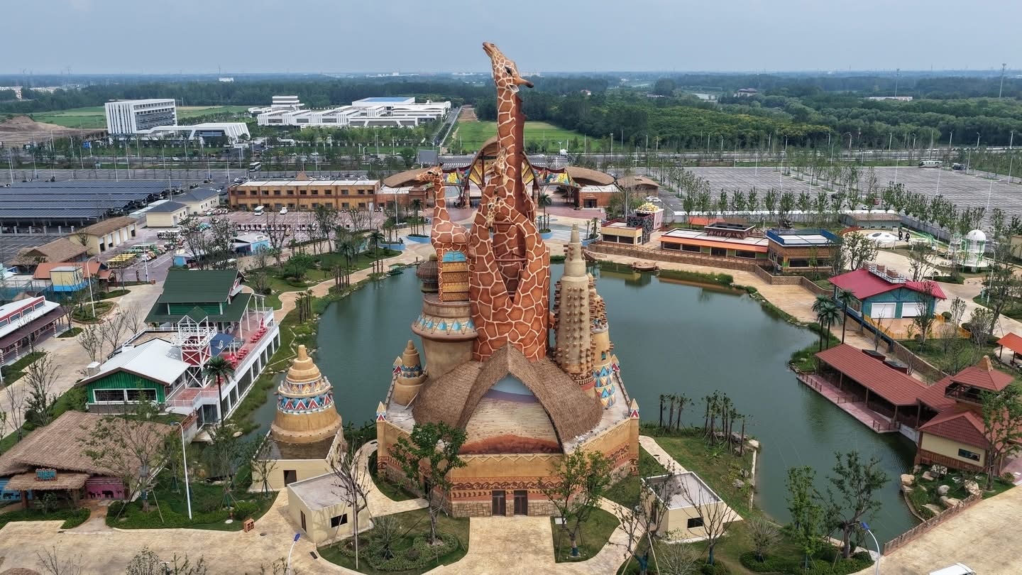 An aerial photograph looking down upon the main entrance plaza of a grand, large-scale safari theme park. The central focus is a massive, intricate wooden and synthetic thatch-roofed gate complex, featuring multiple arches and a prominent central spire. Flanking the front are several other synthetic thatch-roofed buildings serving as ticket offices and shops. A wide, patterned plaza with curving, colored pavement leads to the gate. The park grounds, including a lake, more thatch-roofed structures, dense greenery, and distant city skyscrapers under a gray sky, are visible.