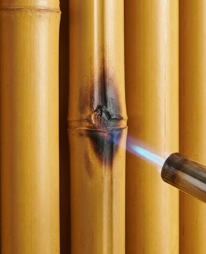 A high-definition macro photograph demonstrating the B1 fire safety of a golden HDPE synthetic bamboo node (reference texture from image_12.png) under precision flame. The material localized scorched (dark brown to black) and shrunk, successfully suppressing the flame without any flaming drips or active burning, proving self-extinguishing performance.
