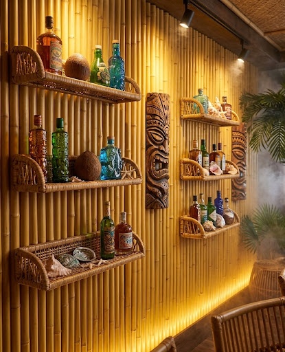 An atmospheric interior photograph of a Tiki restaurant with a seamless background wall covered in golden synthetic bamboo panels, matching image_8.png, serving as a backdrop for shelves filled with vintage rum bottles and carved Tiki masks. Backlighting highlights the bamboo texture.