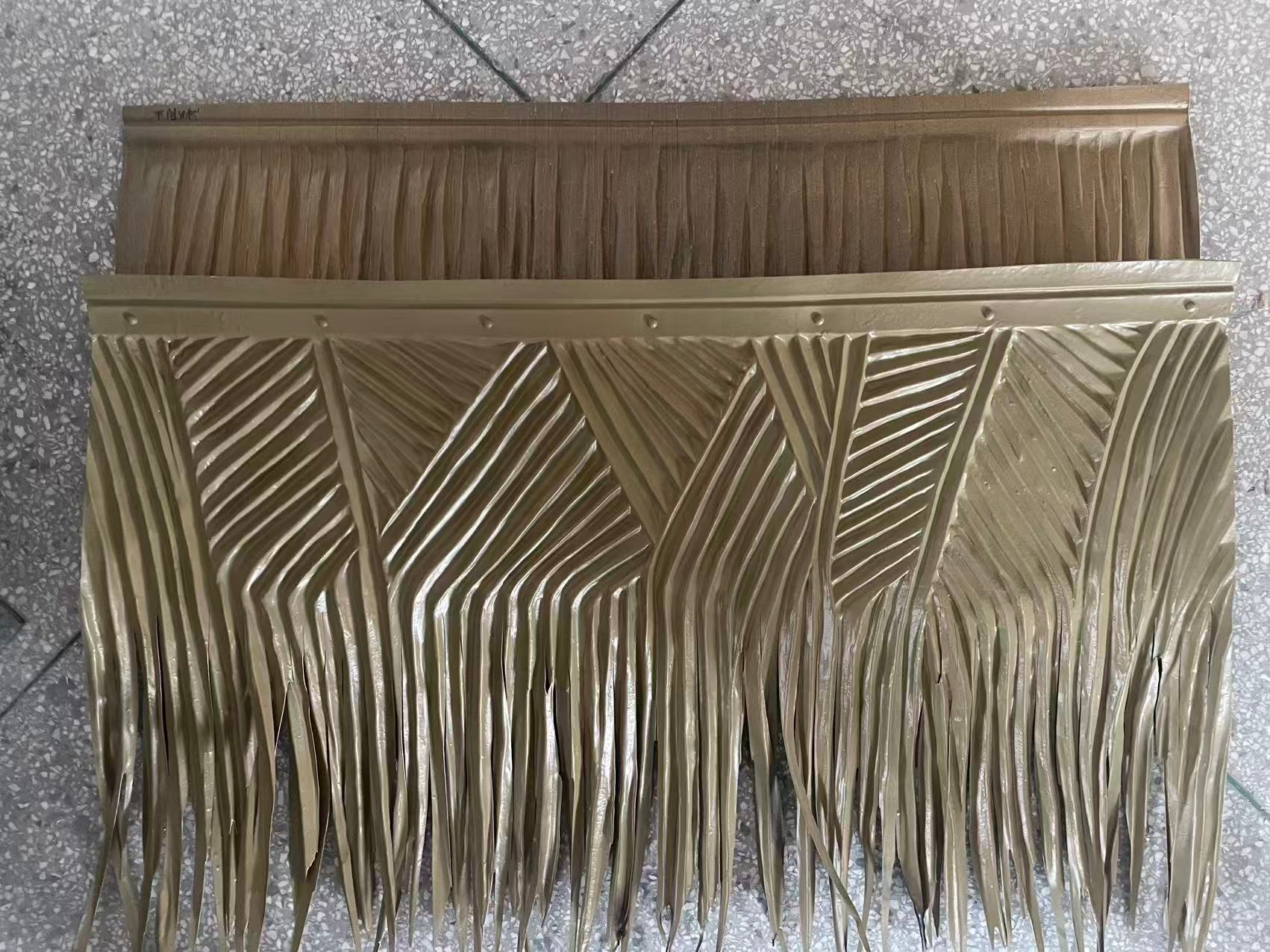 A photograph of two golden-brown Synthetic Nipa thatch panels stacked on a grey speckled floor. The top panel has simple vertical fringing. The bottom panel features an intricate geometric woven pattern with chevrons and diamonds, a studded gold upper border, and fringed bottom edges mimicking traditional natural thatch.